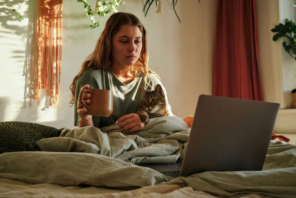 Person sitting in bed holding a mug with a cat in their lap while looking at a laptop in a sunlit bedroom.