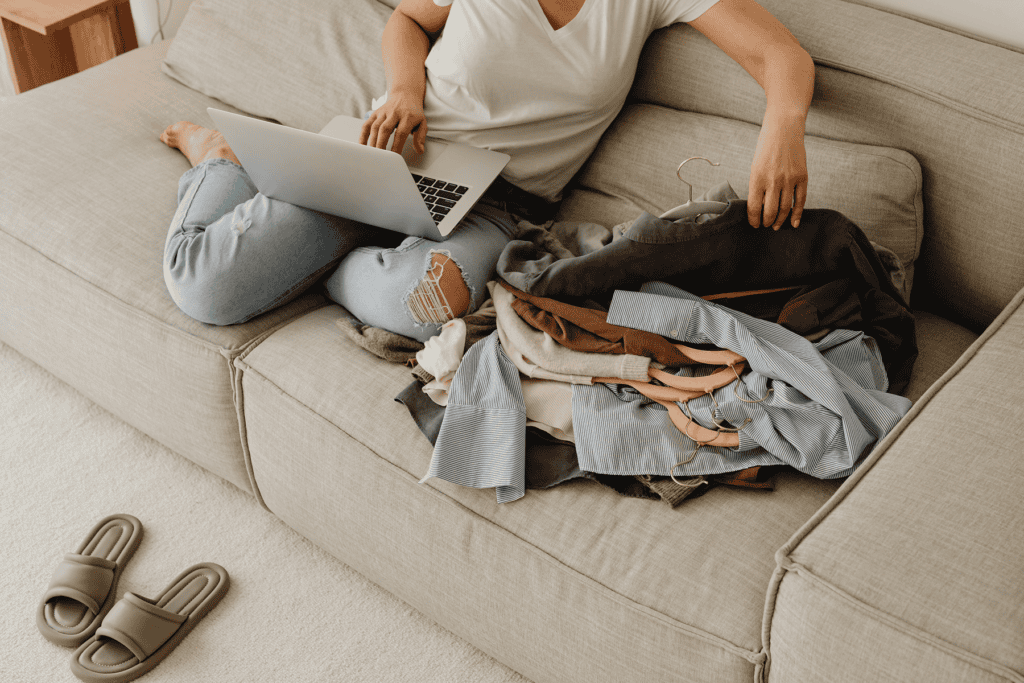 Person sitting on a couch using a laptop while sorting through a pile of clothing on hangers, with more clothes spread across the seat and a pair of slippers on the floor nearby.
