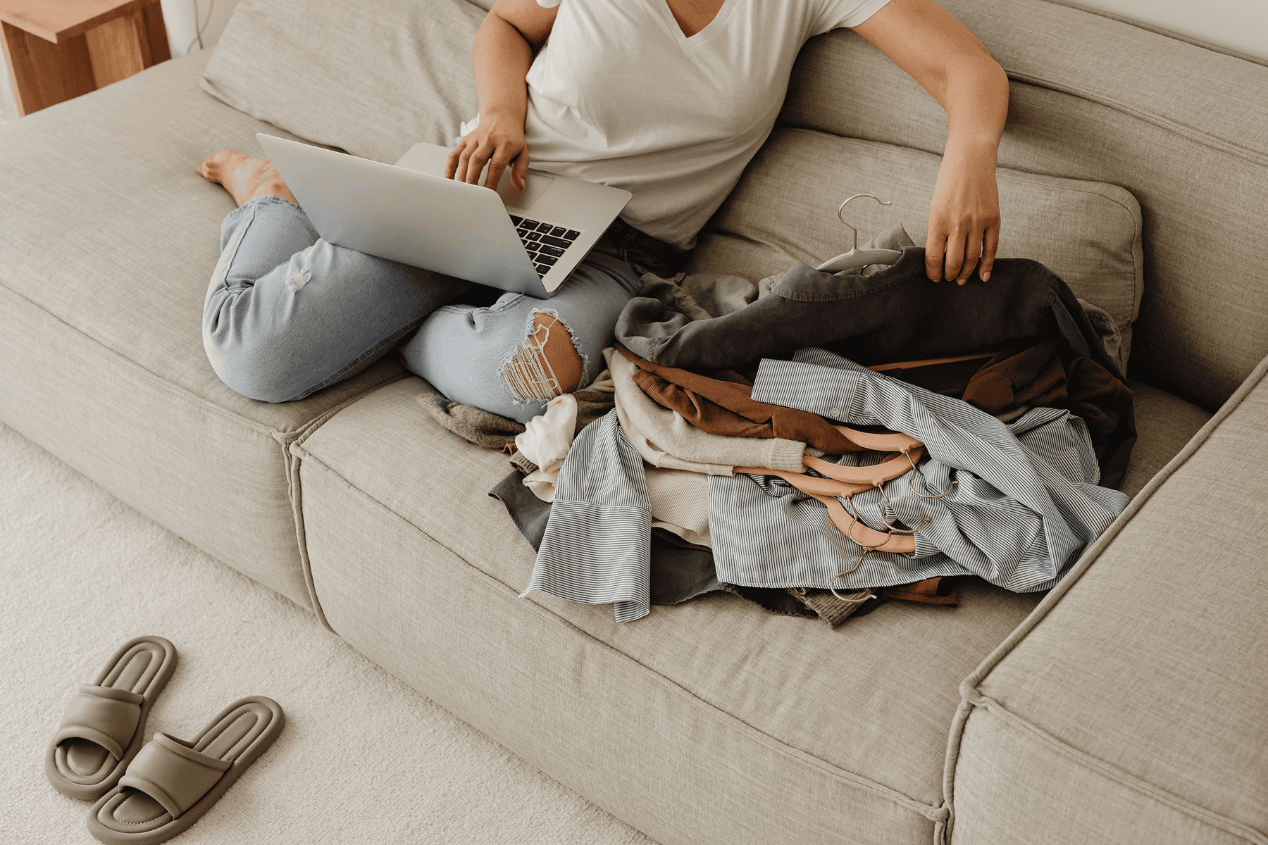 Person sitting on a couch using a laptop while sorting through a pile of clothing on hangers, with more clothes spread across the seat and a pair of slippers on the floor nearby.