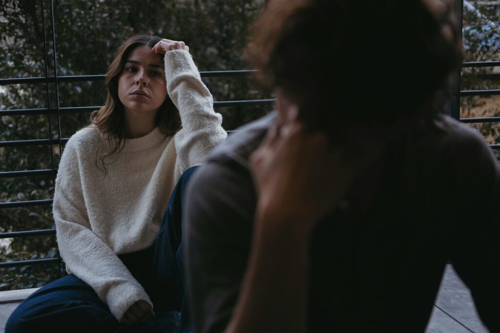 A person sits on the floor near a large window, resting their head on one hand while gazing ahead with a distant, guarded expression. Another person is visible out of focus in the foreground, suggesting emotional distance or a difficult conversation. The scene conveys isolation, emotional withdrawal, or challenges with connection in a relationship.