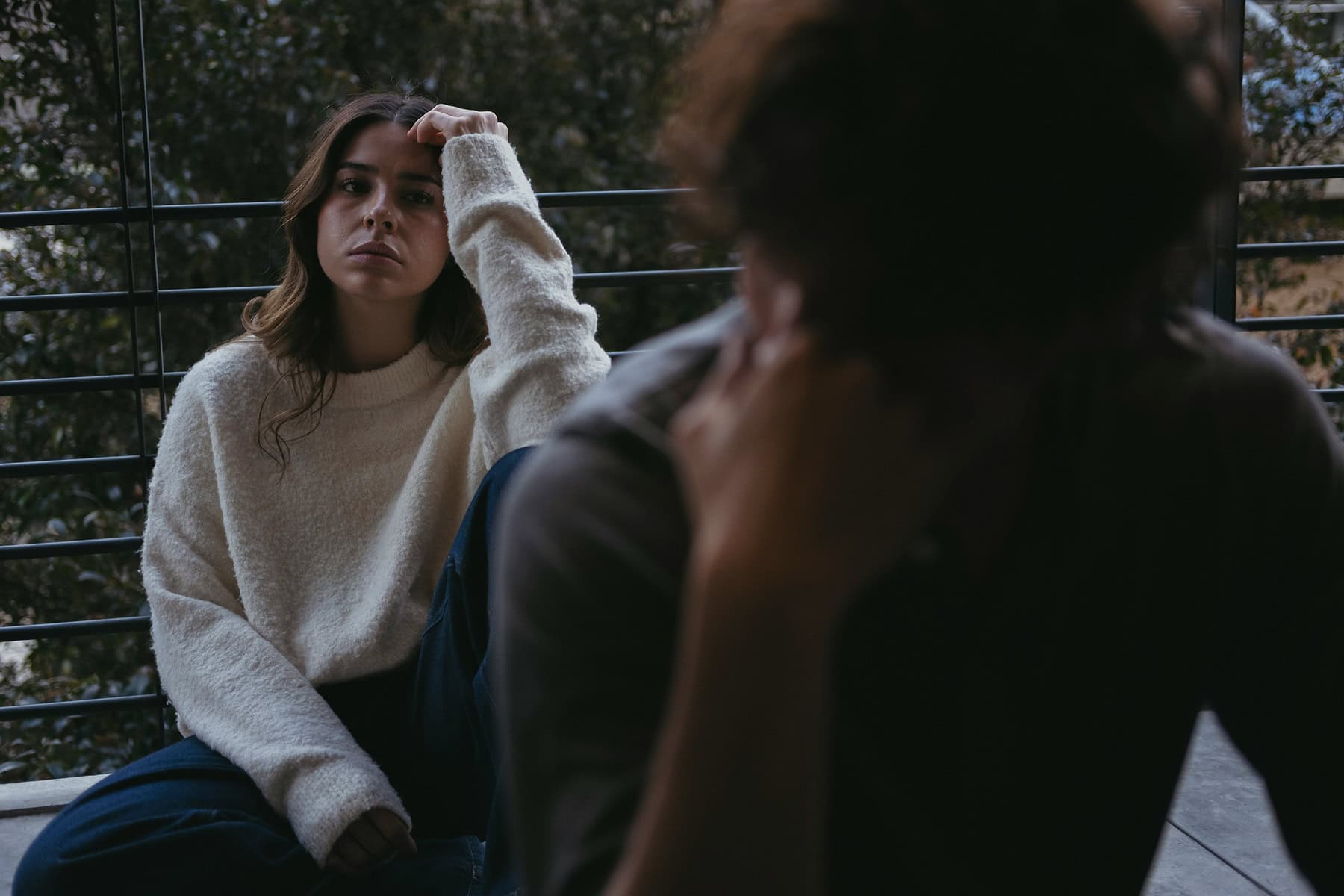 A person sits on the floor near a large window, resting their head on one hand while gazing ahead with a distant, guarded expression. Another person is visible out of focus in the foreground, suggesting emotional distance or a difficult conversation. The scene conveys isolation, emotional withdrawal, or challenges with connection in a relationship.