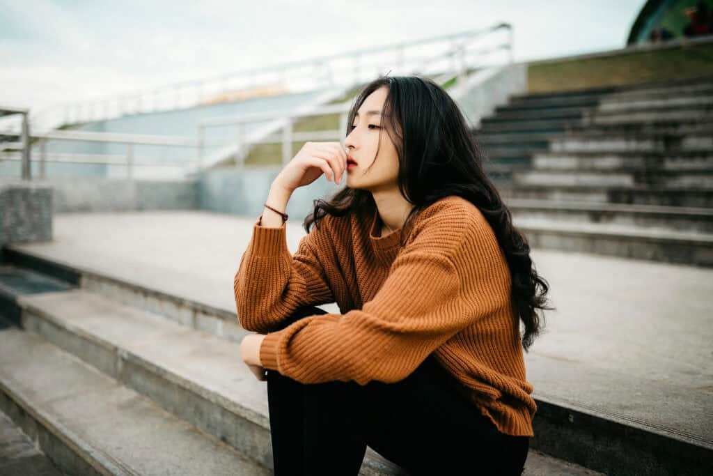 A person sits alone on outdoor concrete steps with one hand resting against their face, looking off into the distance with a thoughtful, subdued expression. The quiet setting and reflective posture convey introspection, worry, or emotional distress.