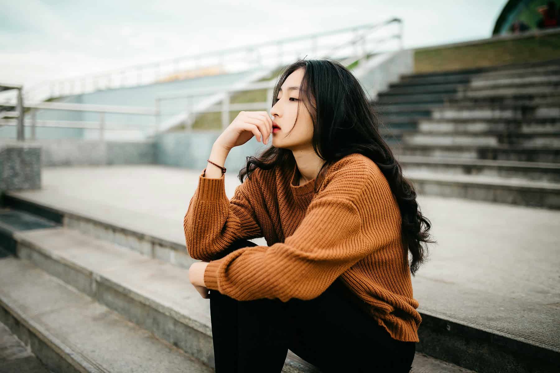A person sits alone on outdoor concrete steps with one hand resting against their face, looking off into the distance with a thoughtful, subdued expression. The quiet setting and reflective posture convey introspection, worry, or emotional distress.