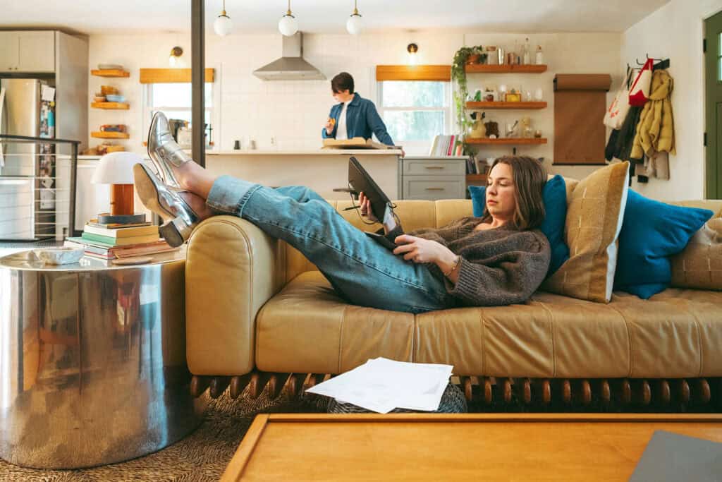 Person reclining on a couch while looking at a tablet and holding a remote, with paperwork on the floor nearby and another person working in the kitchen in the background.