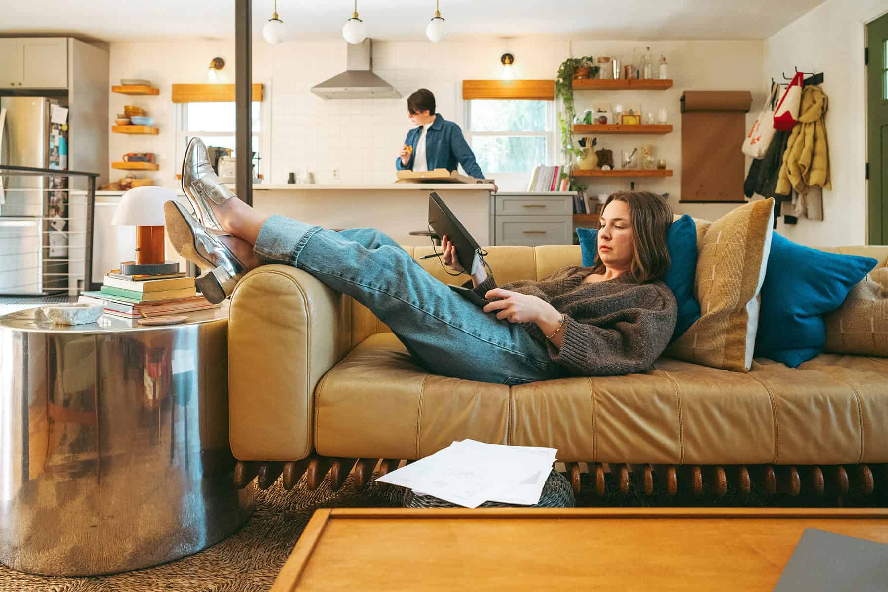 Person reclining on a couch while looking at a tablet and holding a remote, with paperwork on the floor nearby and another person working in the kitchen in the background.