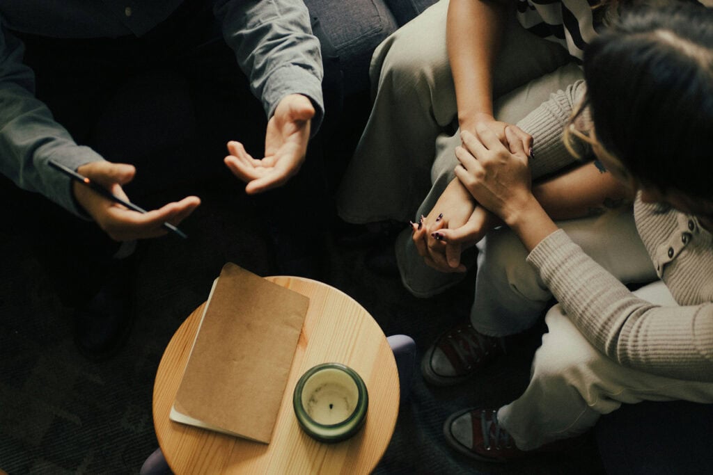 Therapist speaking with a couple during a counseling session as the partners sit close together holding hands, with a notebook and candle on a nearby table.