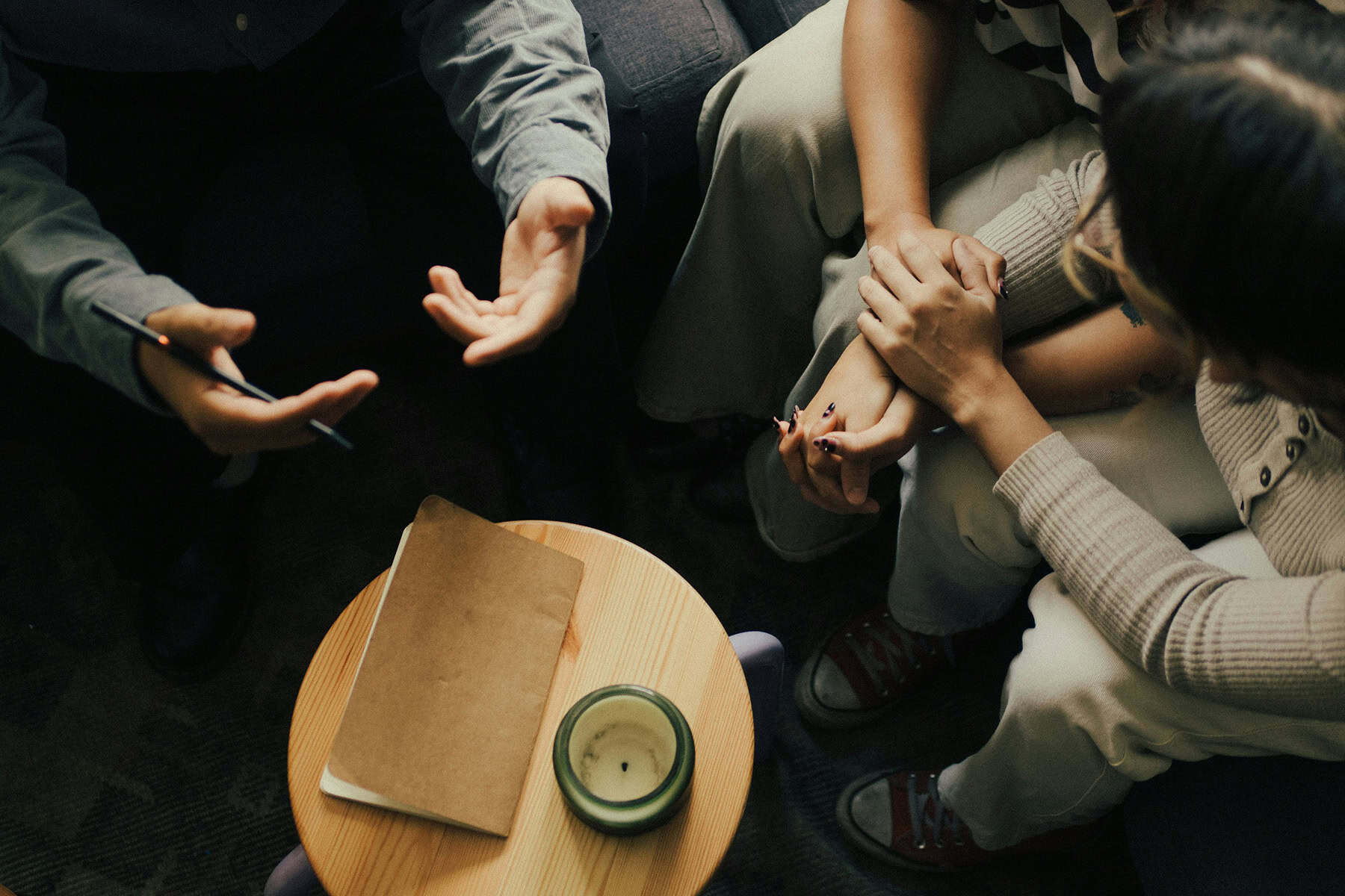 Therapist speaking with a couple during a counseling session as the partners sit close together holding hands, with a notebook and candle on a nearby table.