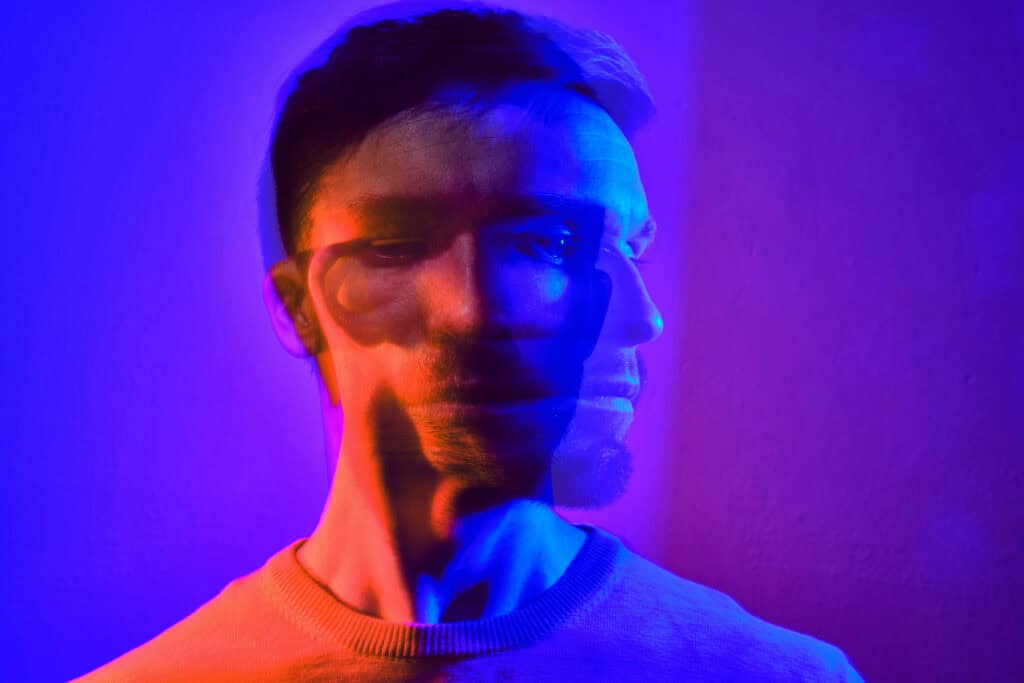 Portrait of a person with a double-exposure effect, creating overlapping facial profiles illuminated by blue, purple, and red lighting.