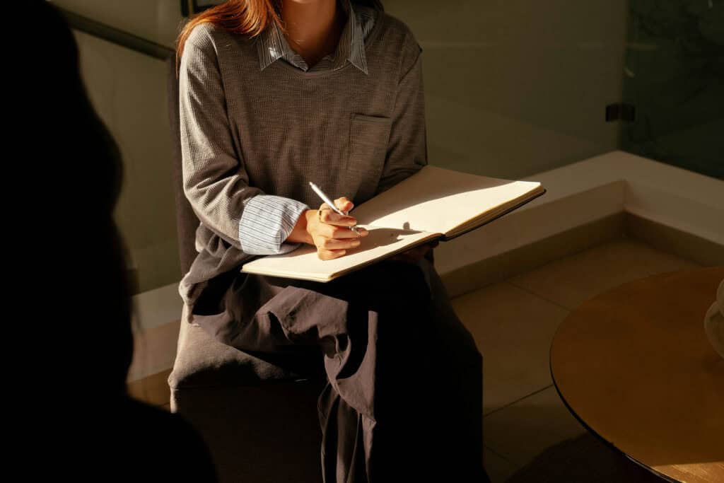 Therapist sitting with a notebook and pen during a counseling session while listening to a client seated nearby.
