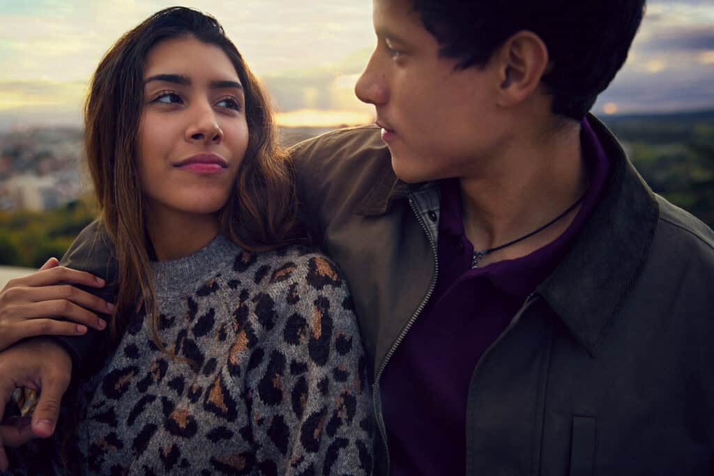 Two people sit close together outdoors at sunset, making eye contact with calm, thoughtful expressions. One person has an arm around the other's shoulders, suggesting closeness and emotional connection, while the blurred landscape in the background creates a quiet, reflective setting.