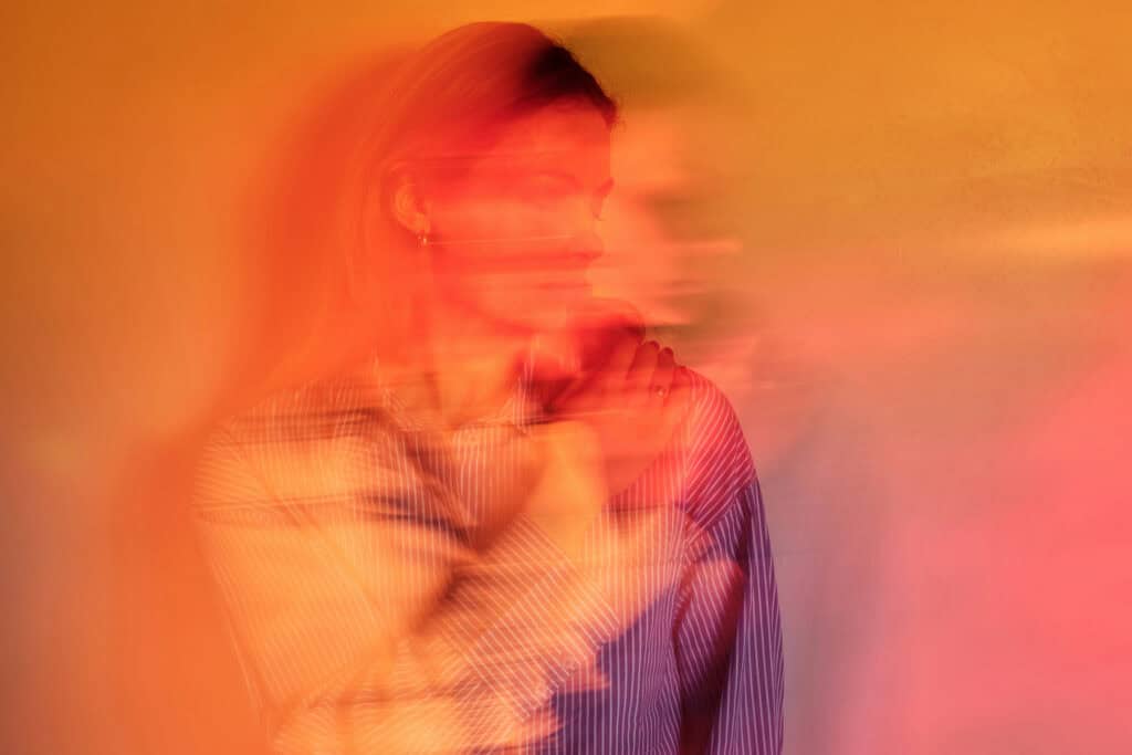 A woman sits with her arms crossed over her chest, her face and body blurred by motion and layered red-orange light, creating a sense of disorientation or detachment.