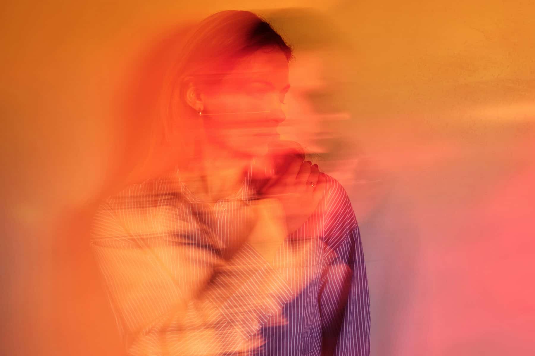 A woman sits with her arms crossed over her chest, her face and body blurred by motion and layered red-orange light, creating a sense of disorientation or detachment.