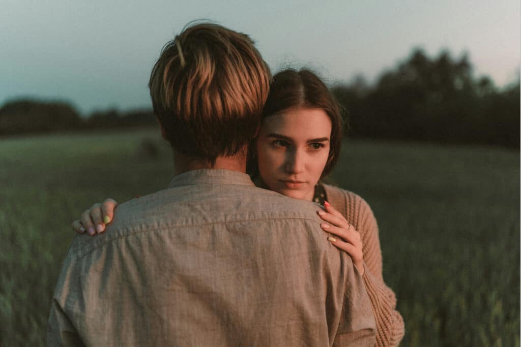 A woman embraces another person outdoors while looking past their shoulder with a serious, distant expression.