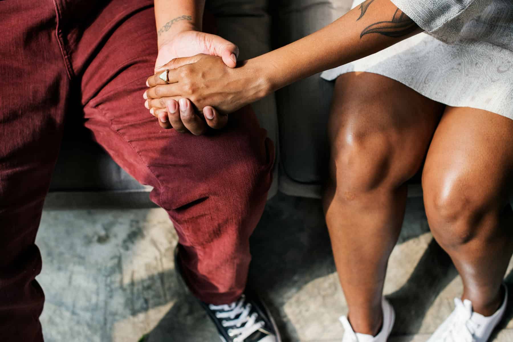 Two people sit closely together, one gently holding the other’s hand with both hands in a supportive, comforting gesture.