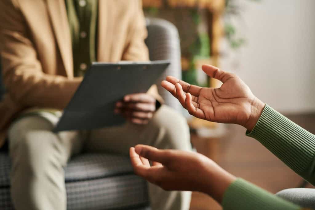 Close-up of a person gesturing with their hands while speaking to a seated therapist holding a clipboard during a counseling session.